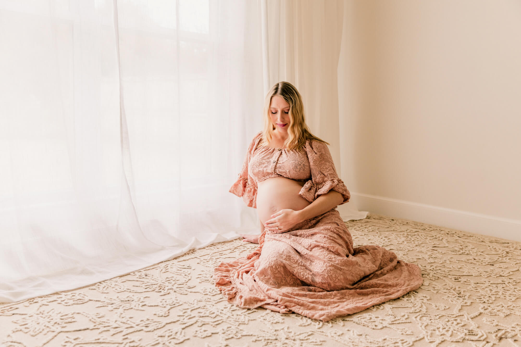 A pregnant lady sits on beige carpet in front of a window while looking down and holding her bump in a pink two piece set.