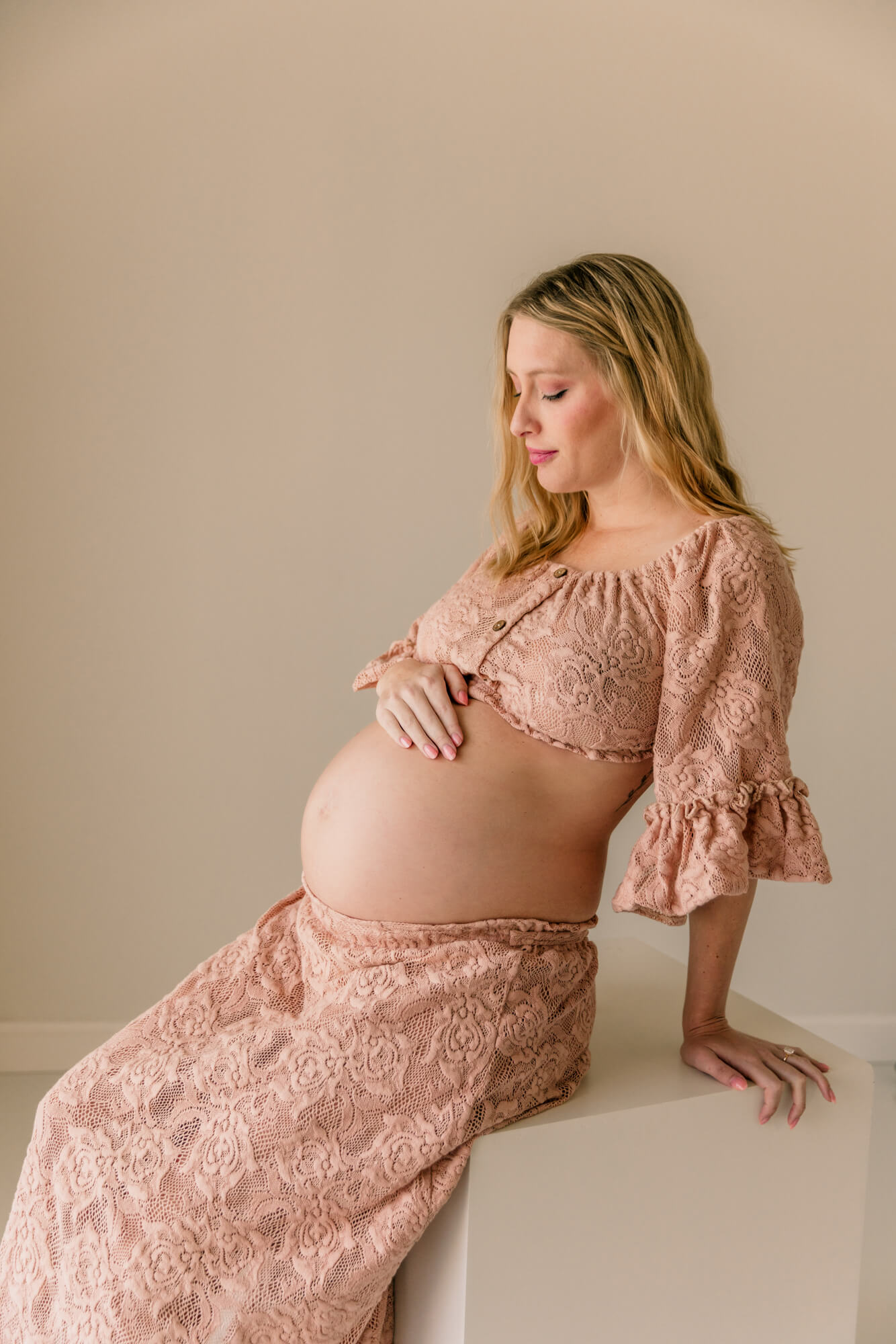 A pregnant lady sits on a white stool with one hand on her bump while wearing a floral laced pink two piece set after browsing OKC maternity stores.