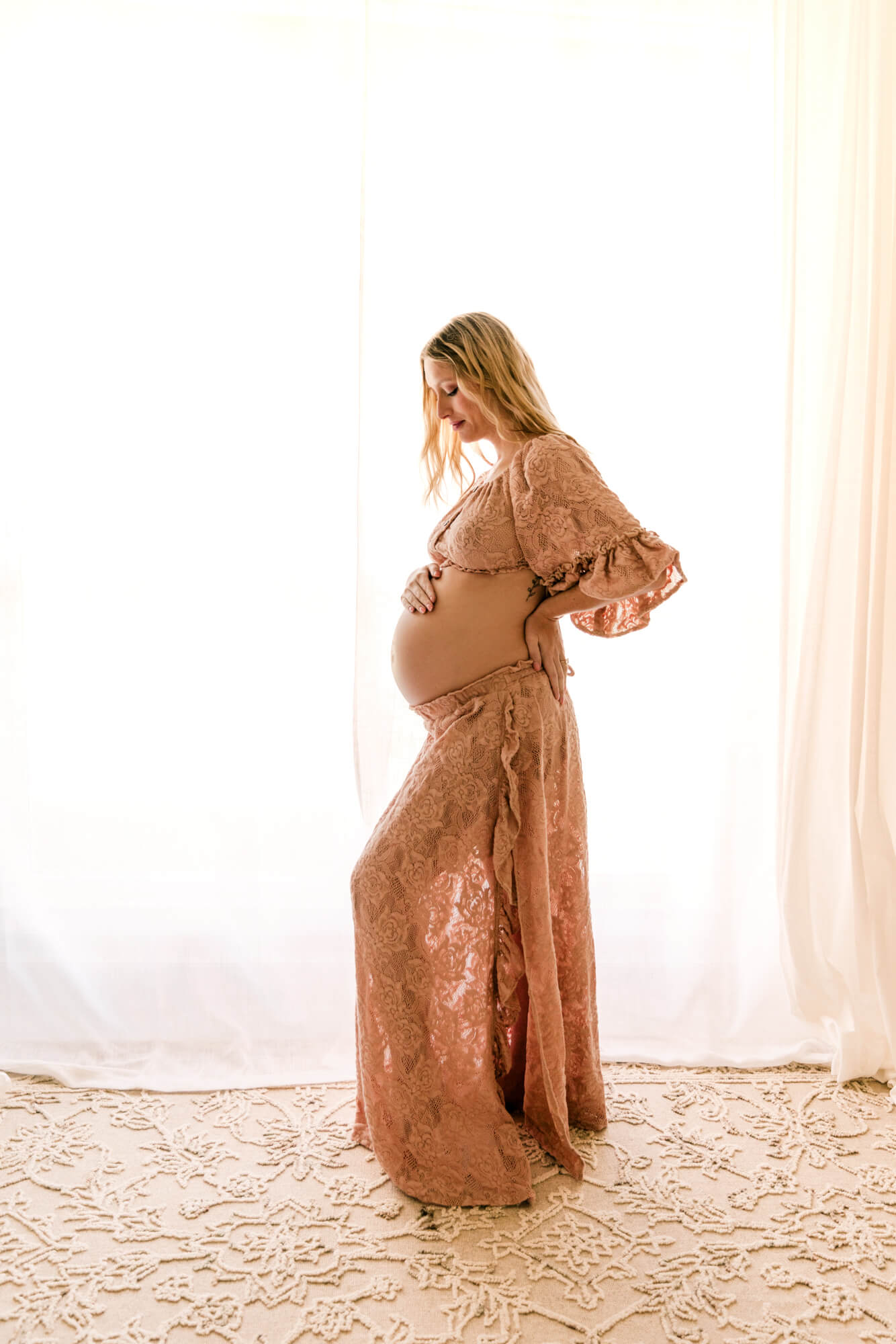 A mother to be in a two piece laced maternity outfit stands in front of a window looking down at her bump with one hand on her bumps and the other on her back after visiting OKC maternity stores.