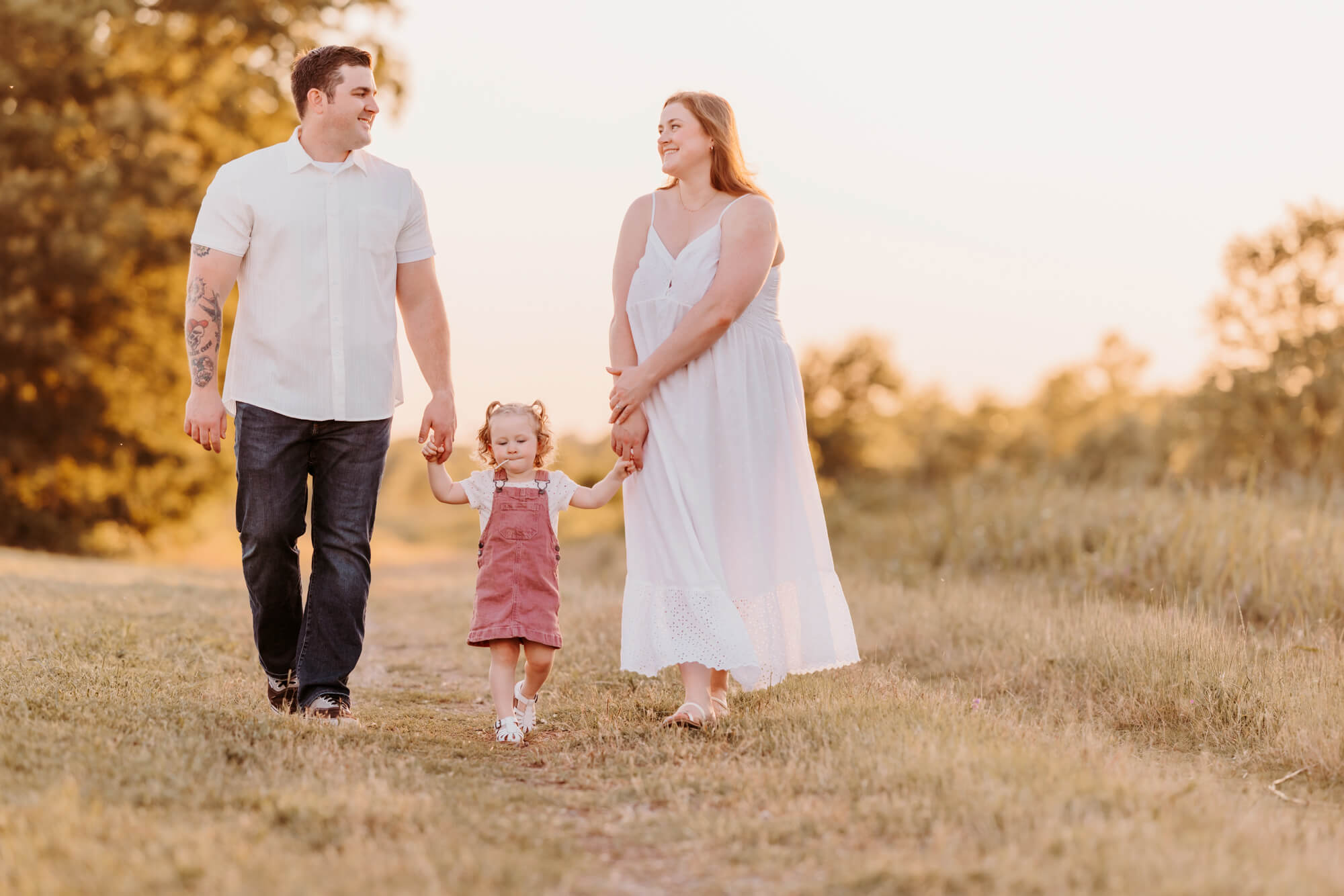 A father and. mother stare at each other and smile while walking in a field of grass holding hands with their young daughter after toddler classes in OKC.