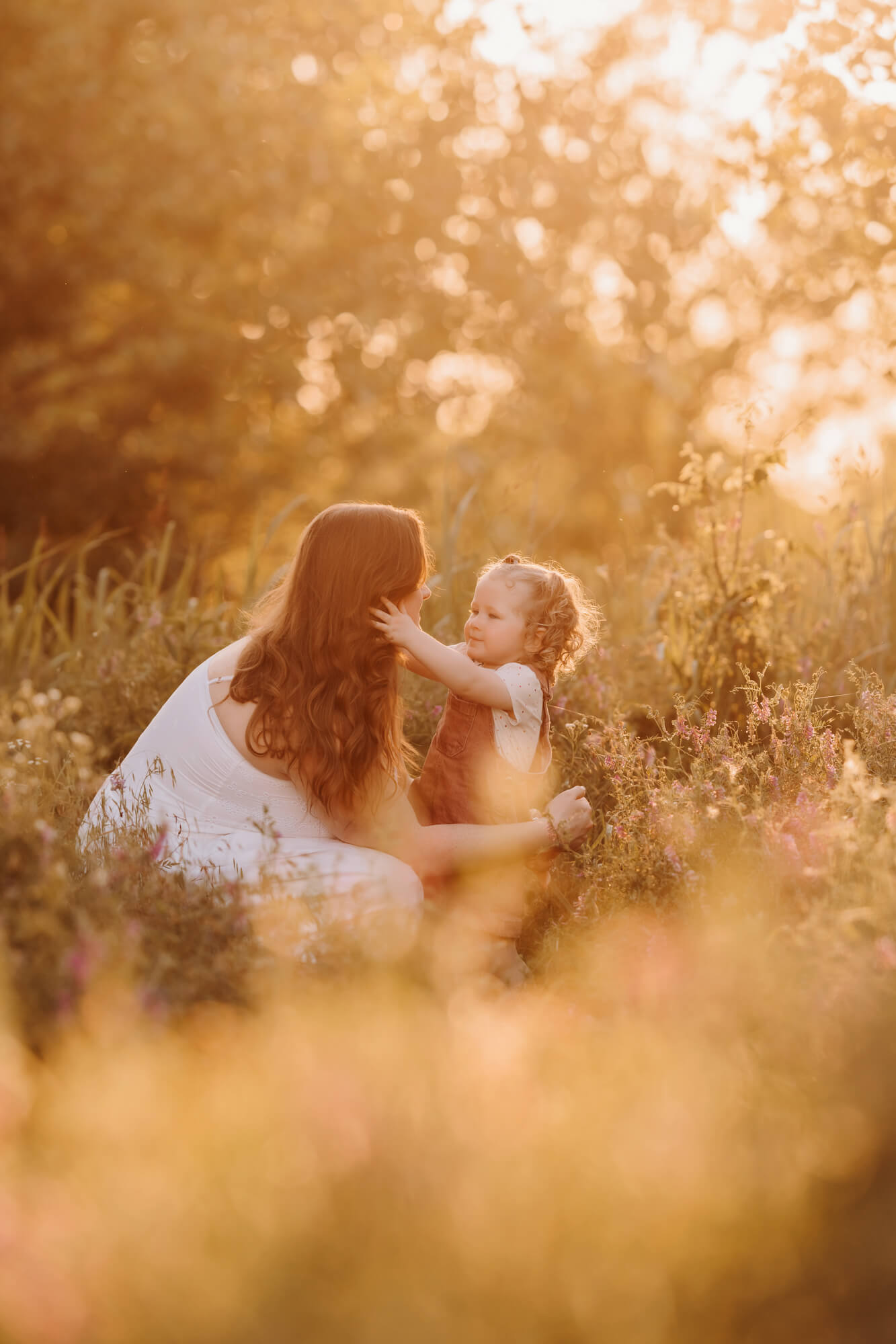 A mom kneels down to her toddler in a field of grass at golden sunset after their toddler classes in OKC.