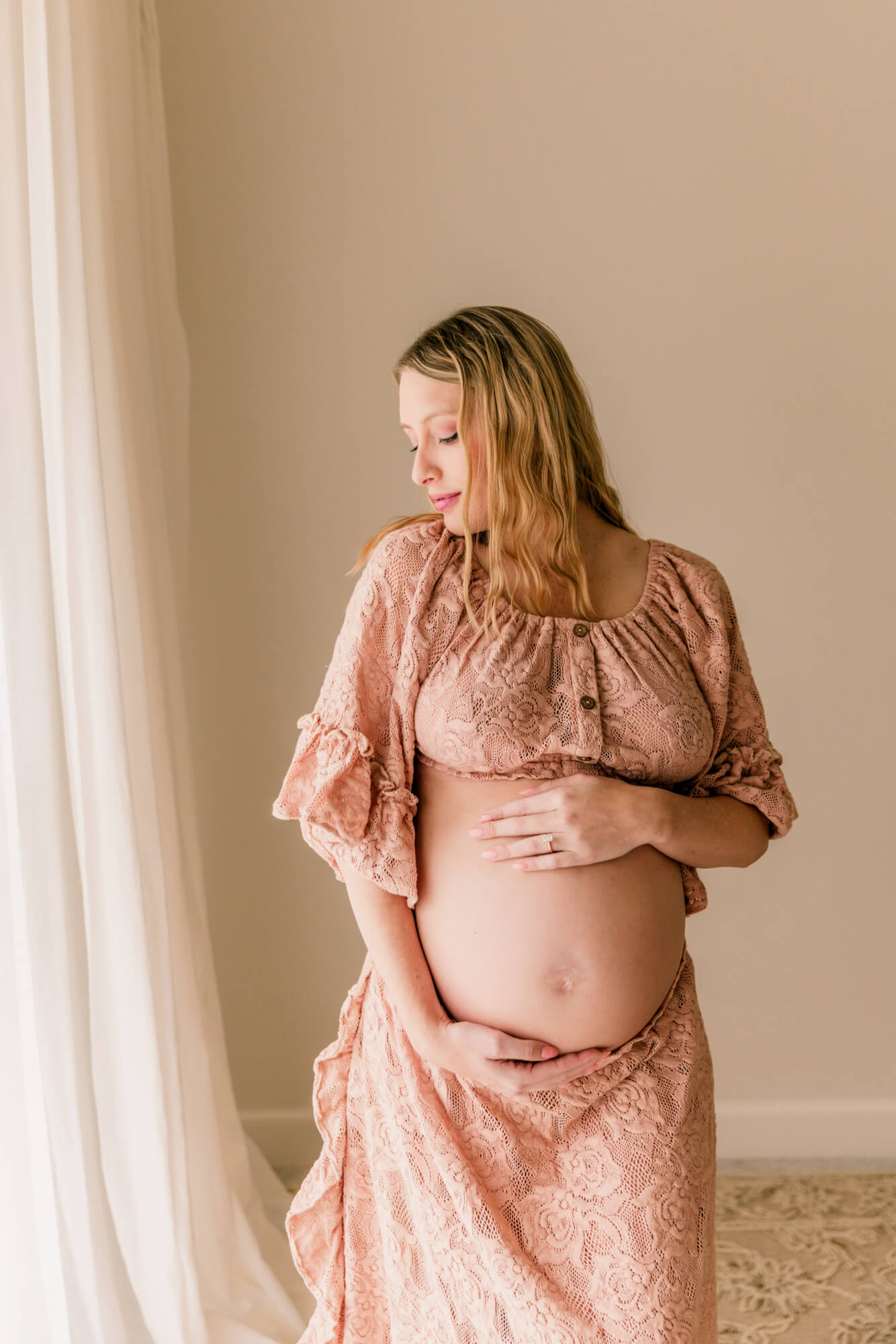 A smiling mother to be gazes down her shoulder holding her bump in a pink lace maternity gown in a studio after a Tulsa prenatal massage