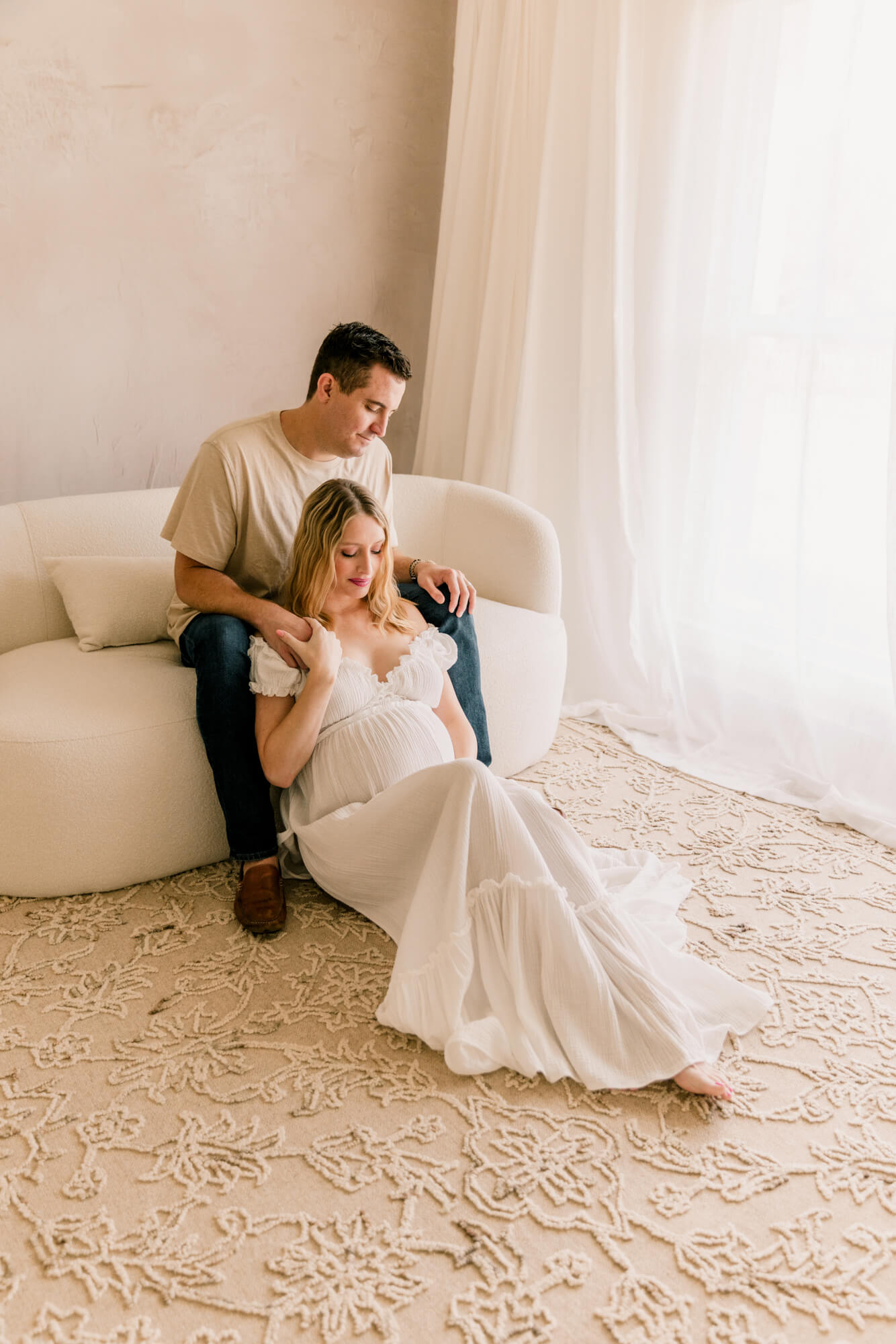 A soon to be dad sits on a beige couch in front of a window with his pregnant wife sitting on the floor leaning against him as they stare down at her bump, after meeting their doula in oklahoma city.