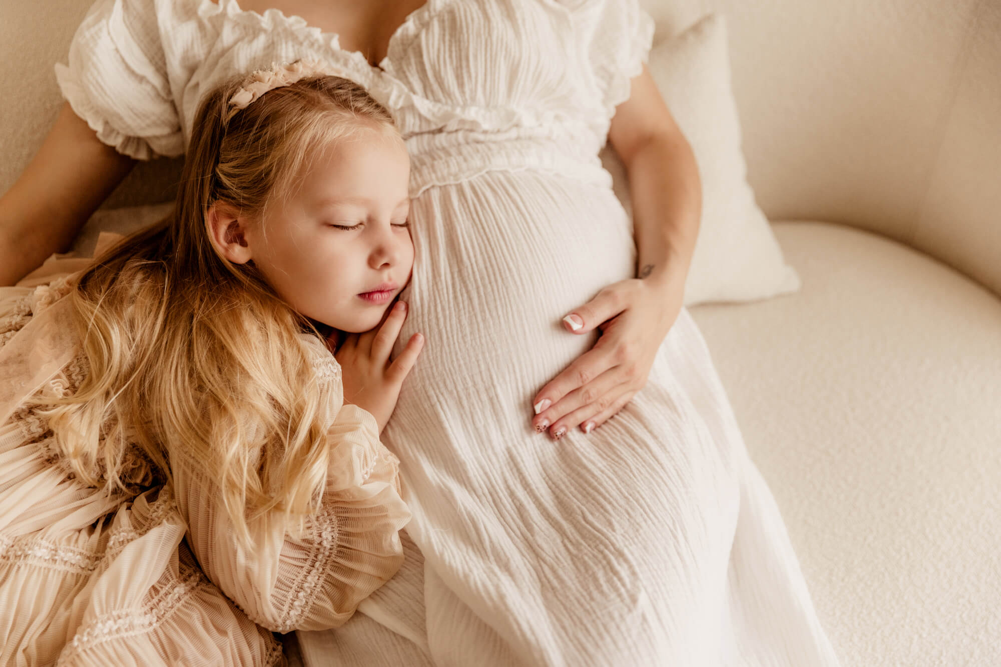 A young girl snuggles against her mom's pregnant belly on a couch in a studio