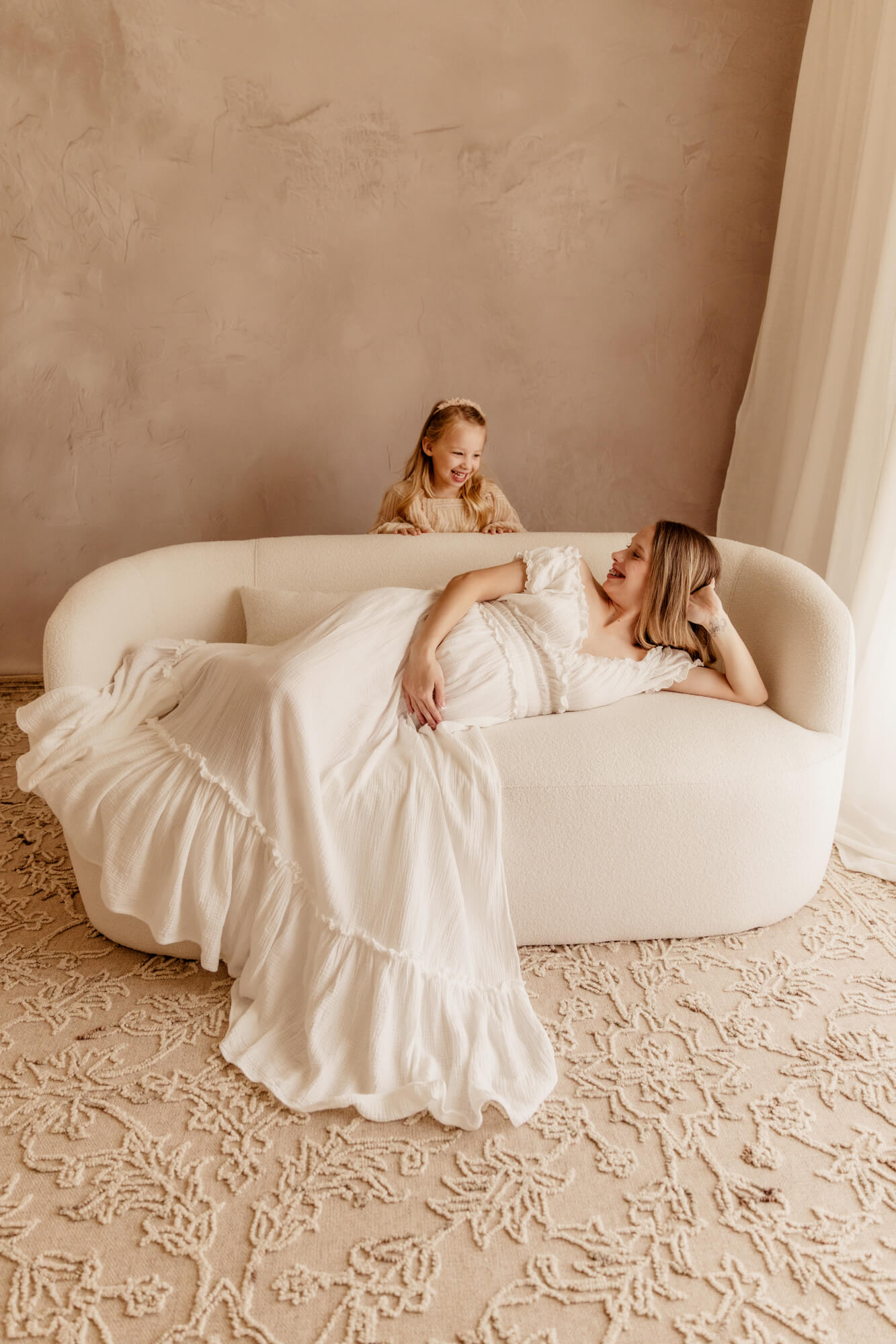 A toddler girl giggles while playing behind a couch her expecting mother is laying across in a studio