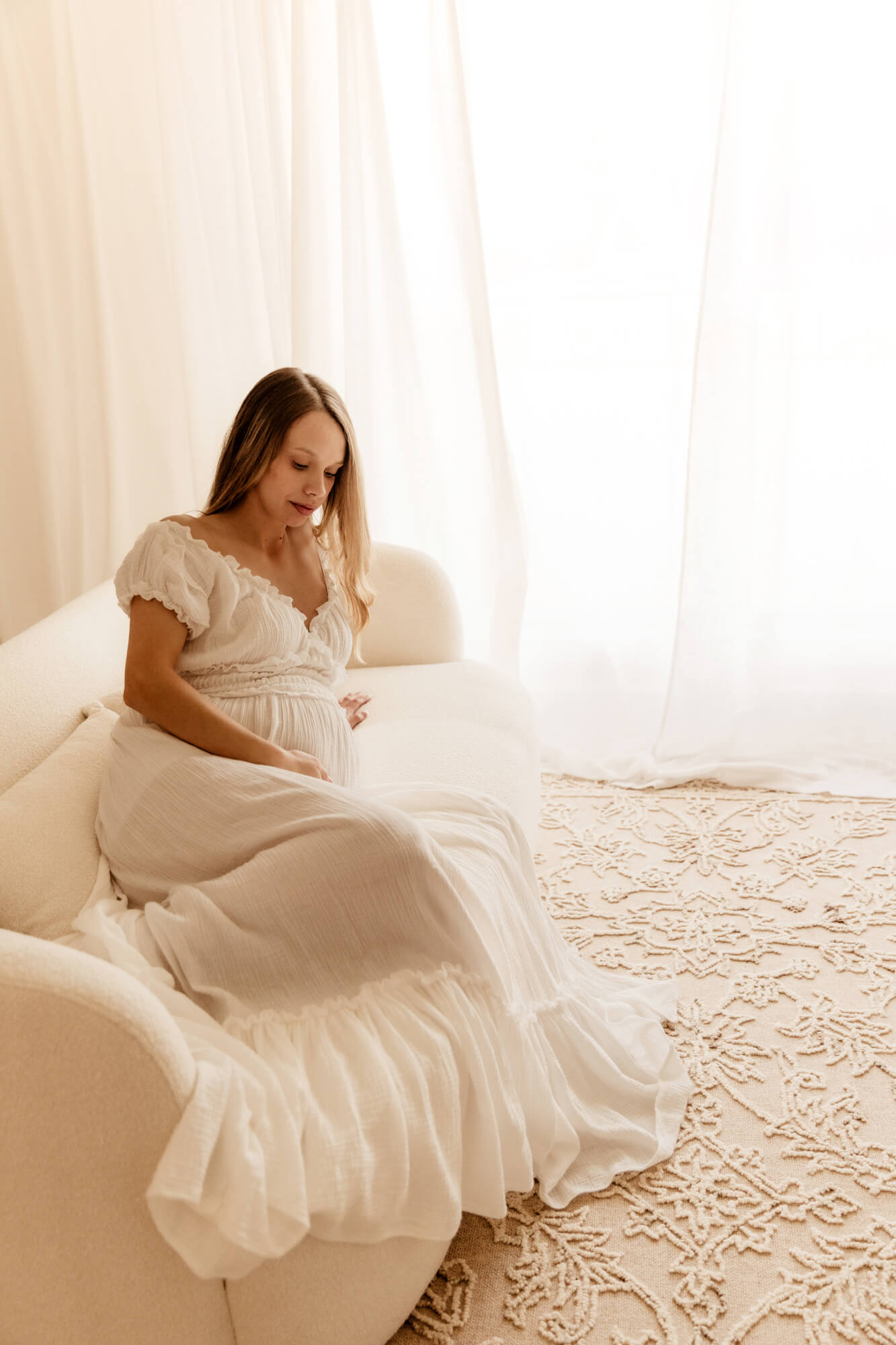 A pregnant mother to be in a white maternity gown lays across a white couch under a large window after visiting a midwife in oklahoma city