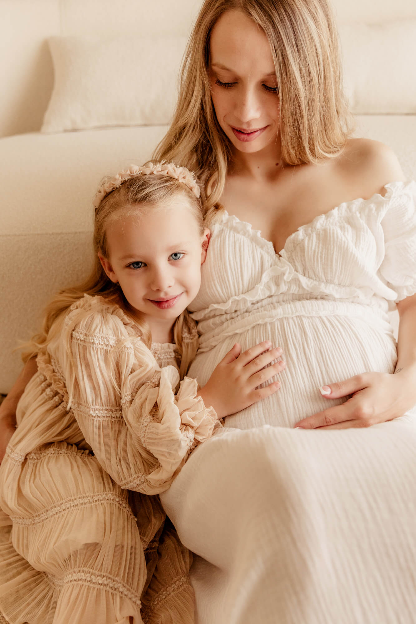 A toddler girl in a cream dress hugs onto mom's pregnant bump while sitting against a couch after meeting a midwife in oklahoma city