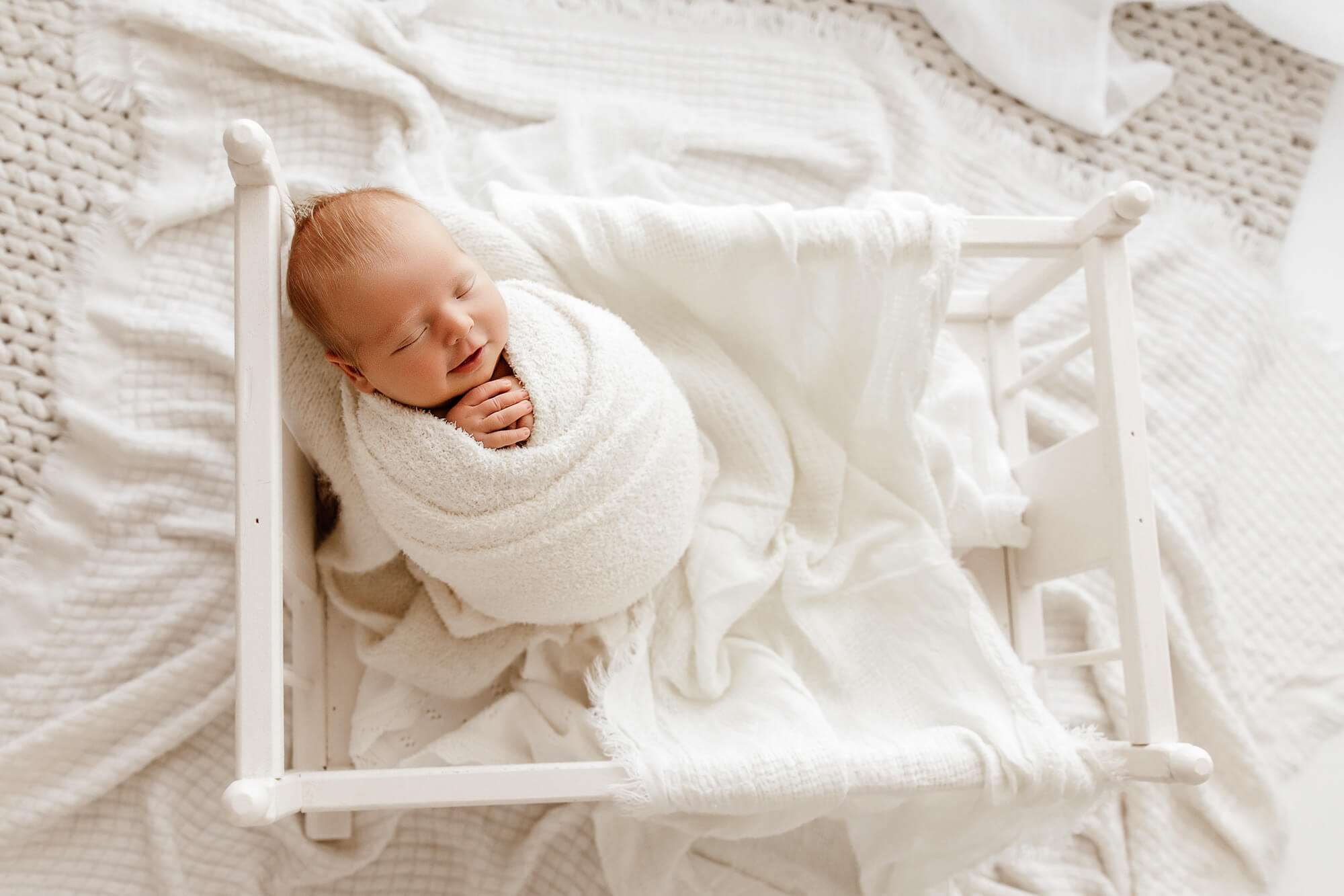 A smiling newborn baby in a white swaddle propped up in a small white crib thanks to a sleep coach in OKC
